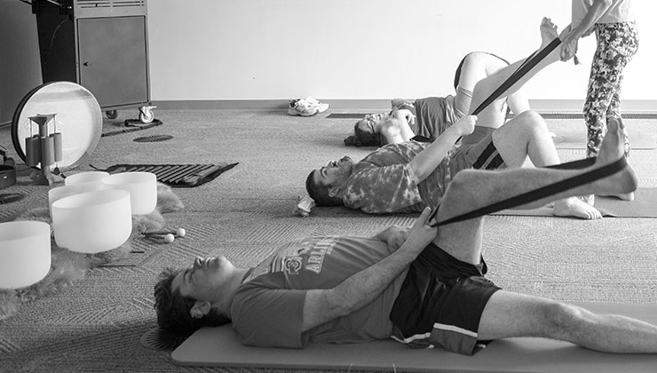 Group practice: participants lying on mats stretching a leg with a yoga strap, crystal singing bowls alongside